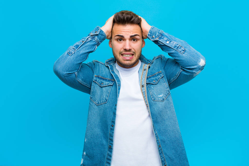 junger hispanischer Mann, der sich gestresst, besorgt, ängstlich oder verängstigt fühlt, mit den Händen auf dem Kopf, vor blauem Hintergrund in Panik - Foto, Bild