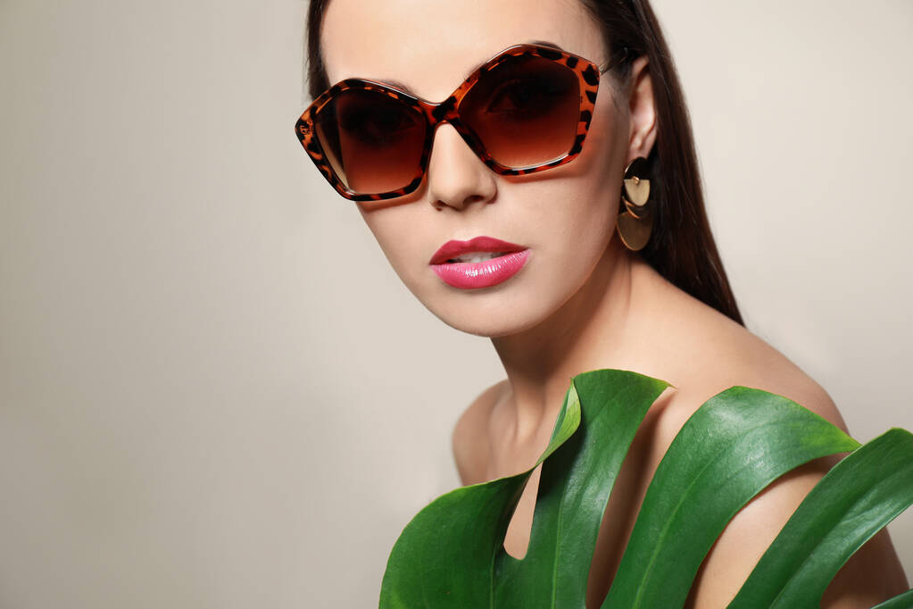 Beautiful woman in stylish sunglasses with tropical leaf on beige background - Photo, Image