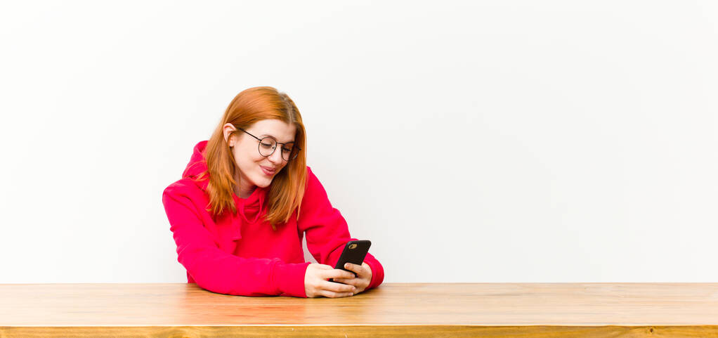 νεαρή κοκκινομάλλα όμορφη γυναίκα μπροστά από ένα ξύλινο τραπέζι με ένα κινητό τηλέφωνο - Φωτογραφία, εικόνα