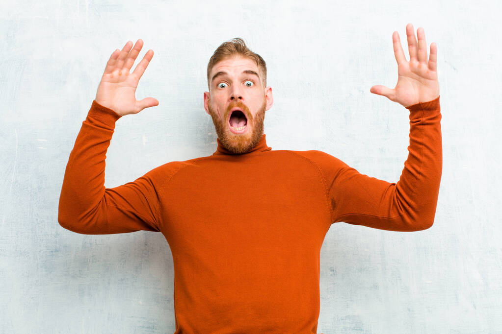 young red head man wearing turtle neck screaming with hands up in the air, feeling furious, frustrated, stressed and upset against cement wall - Photo, Image