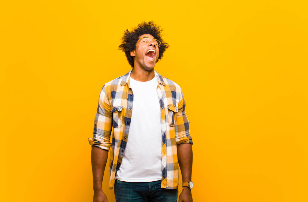 νεαρός μαύρος άντρας να ουρλιάζει μανιωδώς, να φωνάζει επιθετικά, να δείχνει στρεσαρισμένος και θυμωμένος ενάντια στον πορτοκαλί τοίχο - Φωτογραφία, εικόνα