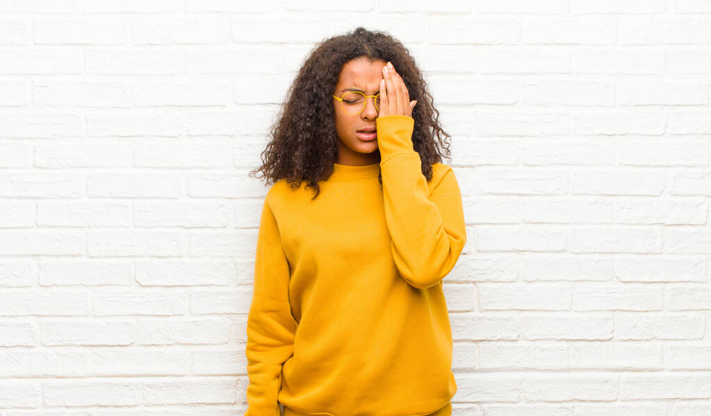 молодая черная женщина выглядит сонной, скучающей и зевающей, с головной болью и одной рукой, прикрывающей половину лица кирпичной стеной
 - Фото, изображение