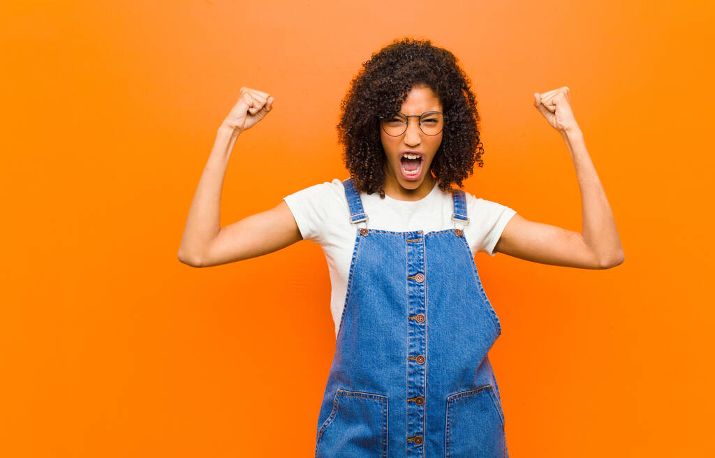 молодая красивая черная женщина, кричащая агрессивно с сердитым выражением лица или с кулаками сжатыми, празднуя успех против оранжевой стены
 - Фото, изображение