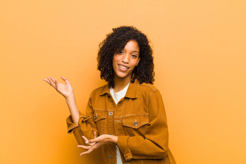 молодая красивая черная женщина улыбается гордо и уверенно, чувствуя себя счастливым и удовлетворенным и показывая концепцию копирования пространства против оранжевой стены
 - Фото, изображение