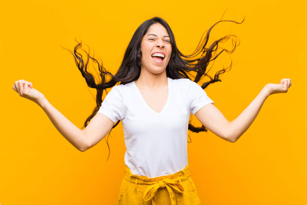 young pretty latin woman looking extremely happy and surprised, celebrating success, shouting and jumping against orange wall - 写真・画像