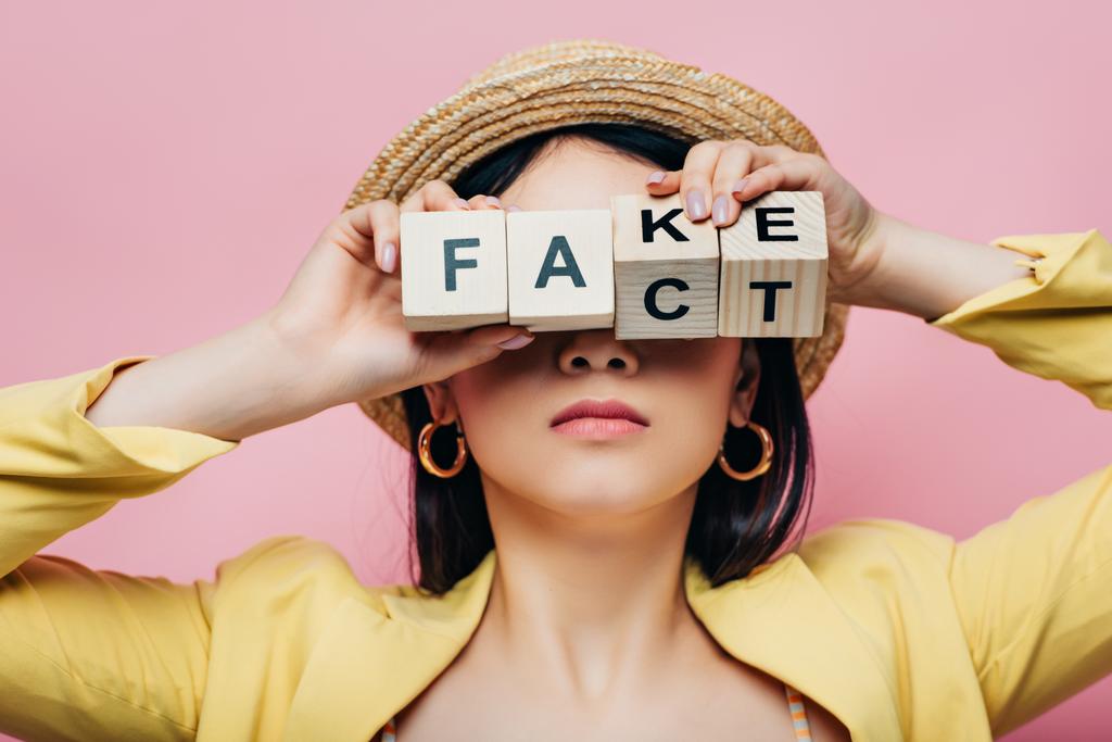 Asijská žena drží dřevěné kostky před obličejem s falešnými a fakta písmo izolované na růžové - Fotografie, Obrázek