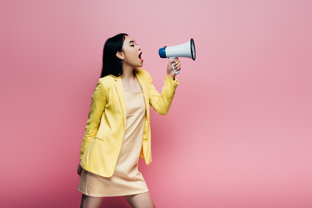 side view of asian woman in yellow outfit screaming in megaphone isolated on pink - Photo, Image