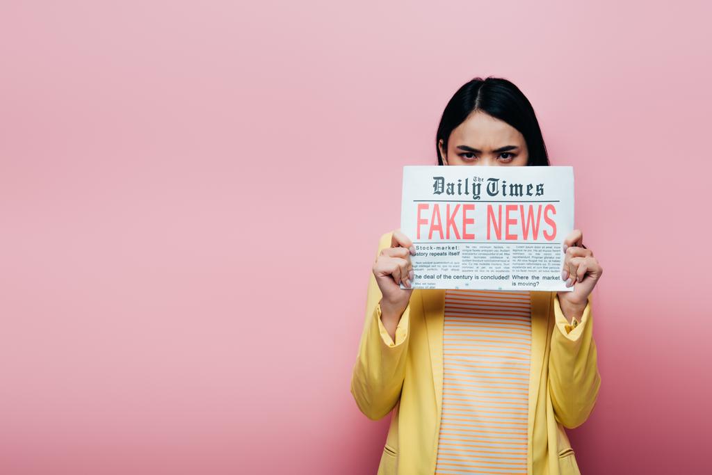 θλιβερή Ασιάτισσα με κίτρινη στολή που κρατάει εφημερίδα με ψεύτικα νέα απομονωμένα στο ροζ. - Φωτογραφία, εικόνα