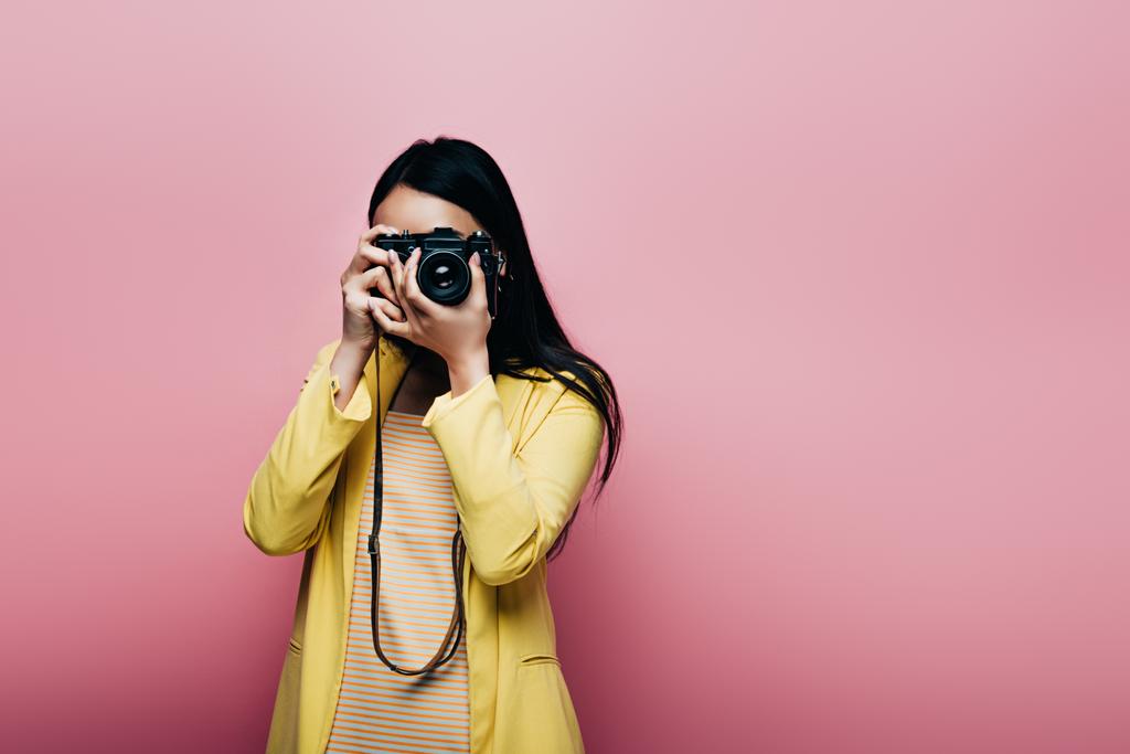 Ασιάτισσα γυναίκα με κίτρινη στολή φωτογραφίζοντας σε ψηφιακή κάμερα απομονωμένη σε ροζ - Φωτογραφία, εικόνα