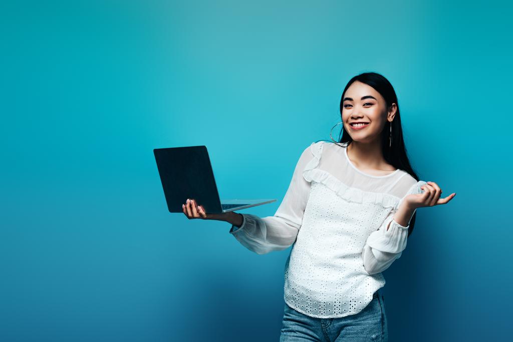 uśmiechnięta azjatycka kobieta w białej bluzce trzyma laptopa na niebieskim tle - Zdjęcie, obraz