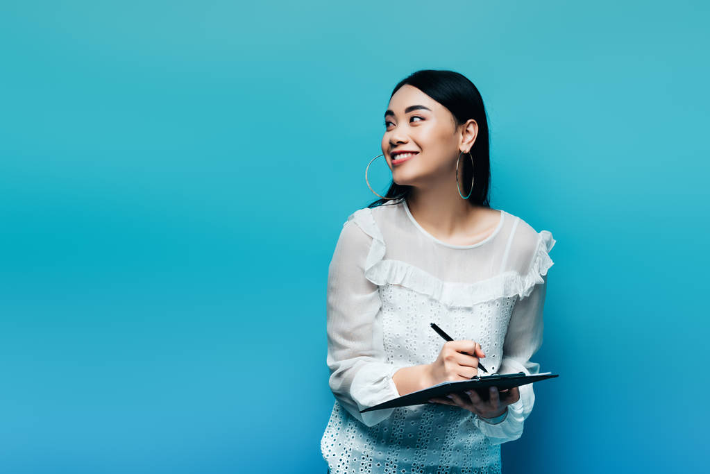 szczęśliwy azjatycki dziennikarz w białej bluzce pisanie na schowku na niebieskim tle - Zdjęcie, obraz