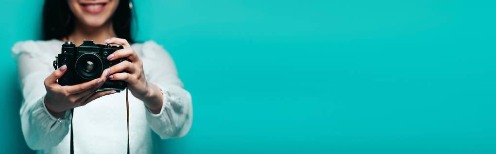przycięty widok uśmiechniętej Azjatki w białej bluzce trzymającej aparat cyfrowy na turkusowym tle, zdjęcie panoramiczne - Zdjęcie, obraz