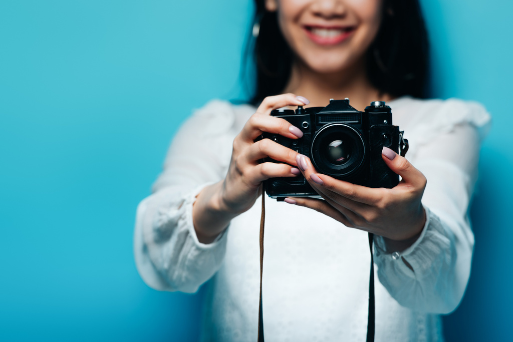 cropped άποψη του χαμογελαστού ασιατική γυναίκα σε λευκό μπλούζα κρατώντας ψηφιακή φωτογραφική μηχανή σε μπλε φόντο - Φωτογραφία, εικόνα