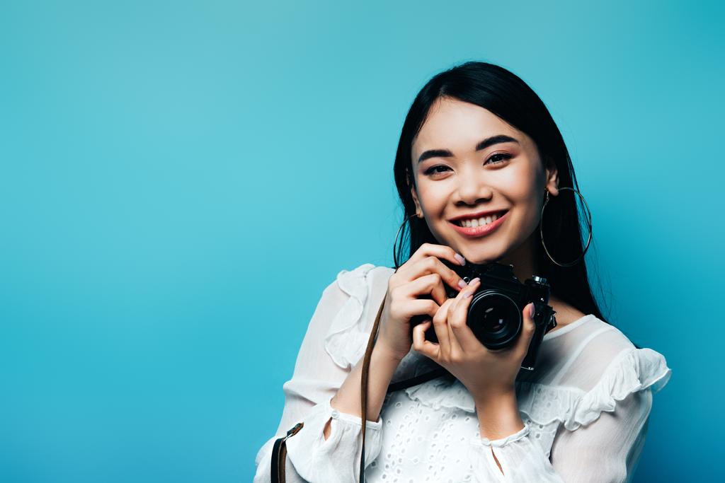 щаслива азіатська жінка в білій блузці тримає цифрову камеру на синьому фоні
 - Фото, зображення