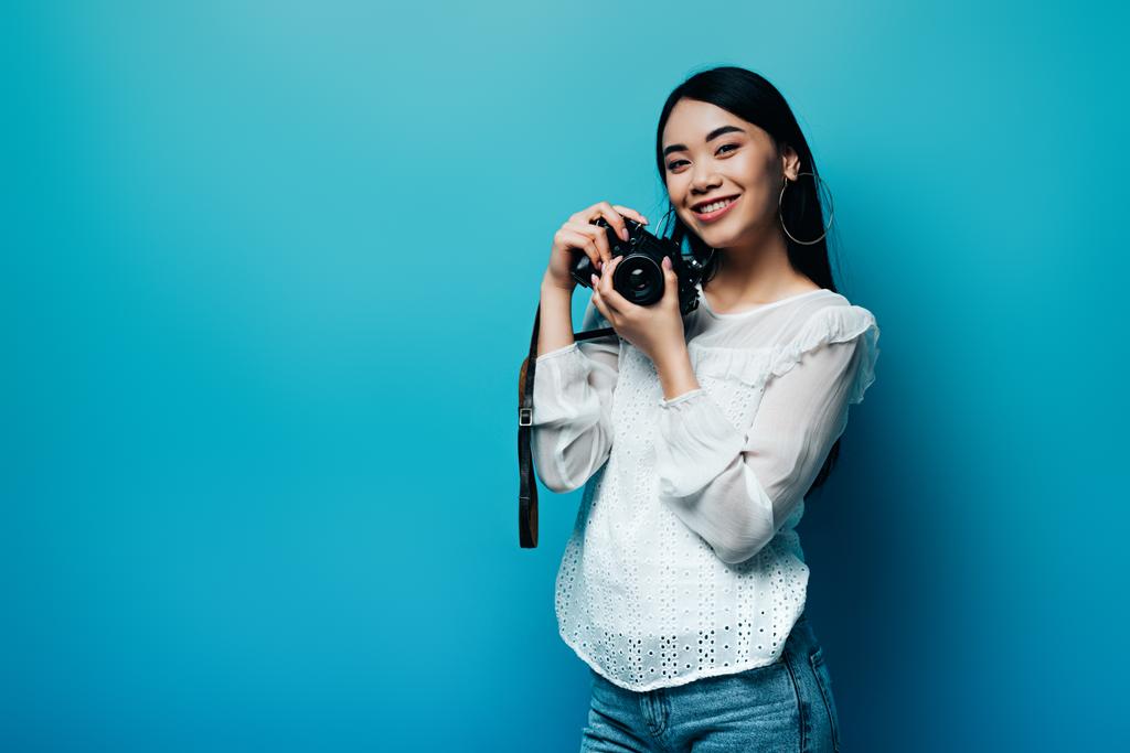 happy asian woman in white blouse holding digital camera on blue background - Photo, Image