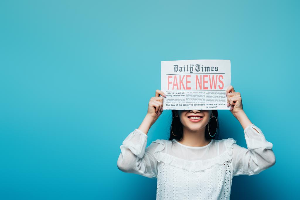 smiling asian woman in white blouse holding newspaper with fake news on blue background - Photo, Image