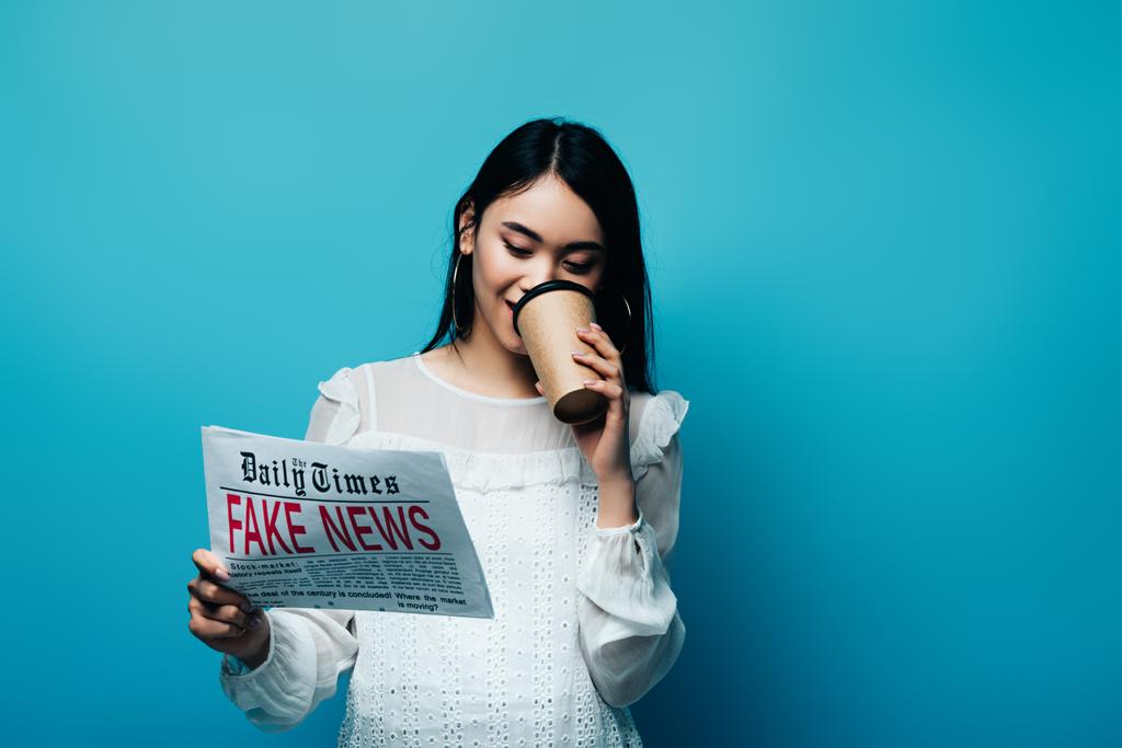 mujer asiática en blusa blanca sosteniendo periódico con noticias falsas y bebiendo café sobre fondo azul
 - Foto, Imagen