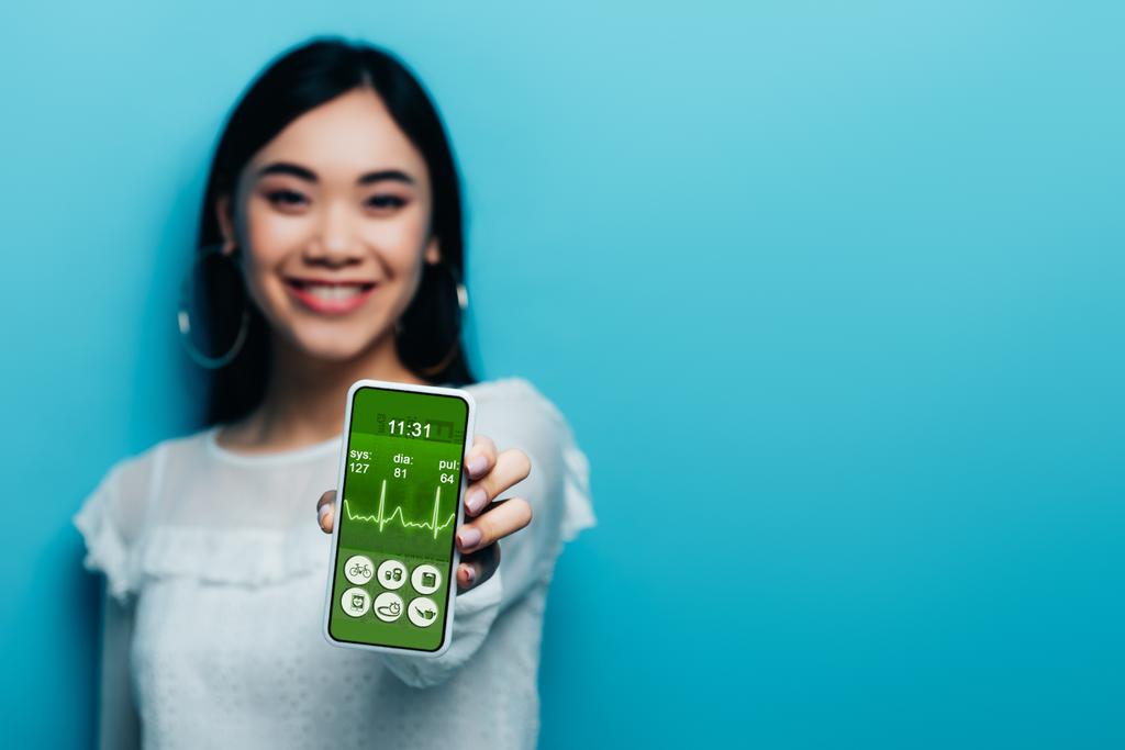 Selektiver Fokus einer lächelnden asiatischen Frau in weißer Bluse mit Smartphone mit Gesundheits-App auf blauem Hintergrund - Foto, Bild