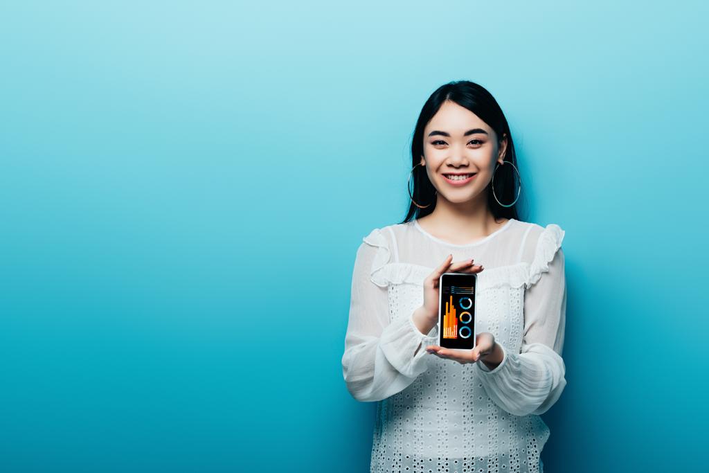 улыбающаяся азиатка в белой блузке, держащая смартфон с бизнес-приложением на синем фоне
 - Фото, изображение