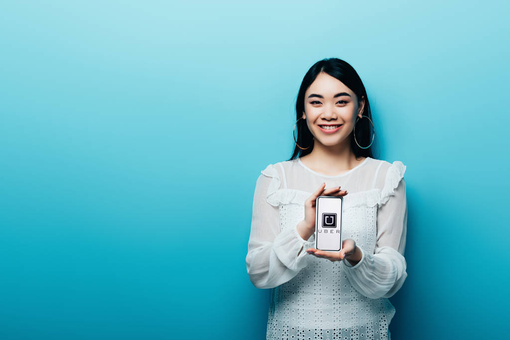 Kiev, Oekraïne - 15 juli 2019: lachende Aziatische vrouw in witte blouse met smartphone met uber app op blauwe achtergrond - Foto, afbeelding