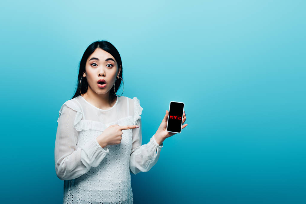 Київ, Україна - 15 липня 2019: шокована жінка-азіат, що вказує пальцем на смартфон з застосунком netflix на синьому фоні. - Фото, зображення