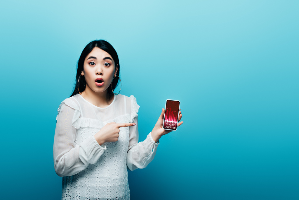 conmocionado mujer asiática señalando con el dedo en el teléfono inteligente con cursos de comercio aplicación sobre fondo azul
 - Foto, Imagen