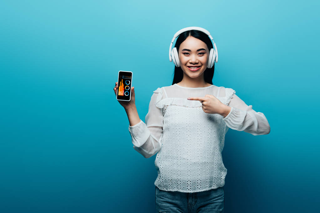 smiling asian woman with headphones pointing with finger at smartphone with business app on blue background - Photo, Image
