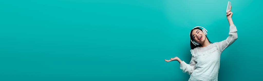 sorrindo mulher asiática com olhos fechados em fones de ouvido segurando smartphone e dançando em fundo turquesa, tiro panorâmico
 - Foto, Imagem