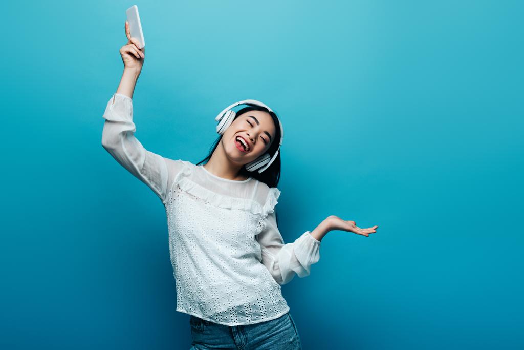 χαμογελαστή Ασιάτισσα με κλειστά μάτια στα ακουστικά κρατώντας smartphone και χορεύοντας σε μπλε φόντο - Φωτογραφία, εικόνα