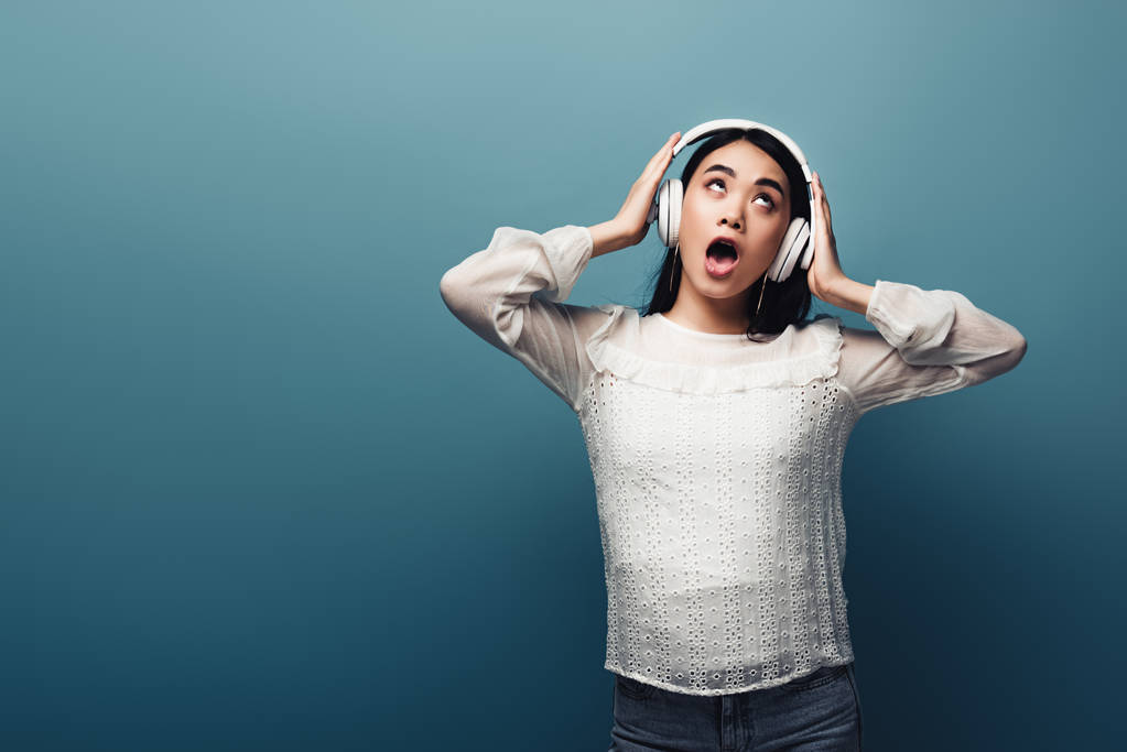 schockierte asiatische Frau mit Händen auf Kopfhörern und offenem Mund auf blauem Hintergrund - Foto, Bild