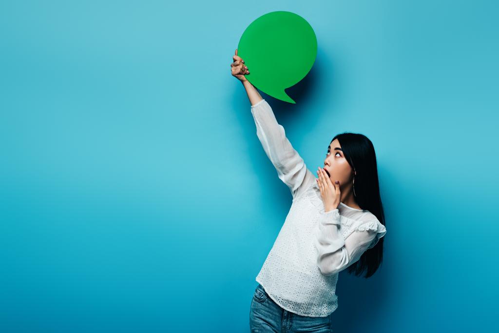 shocked brunette asian woman holding green speech bubble on blue background - Photo, Image