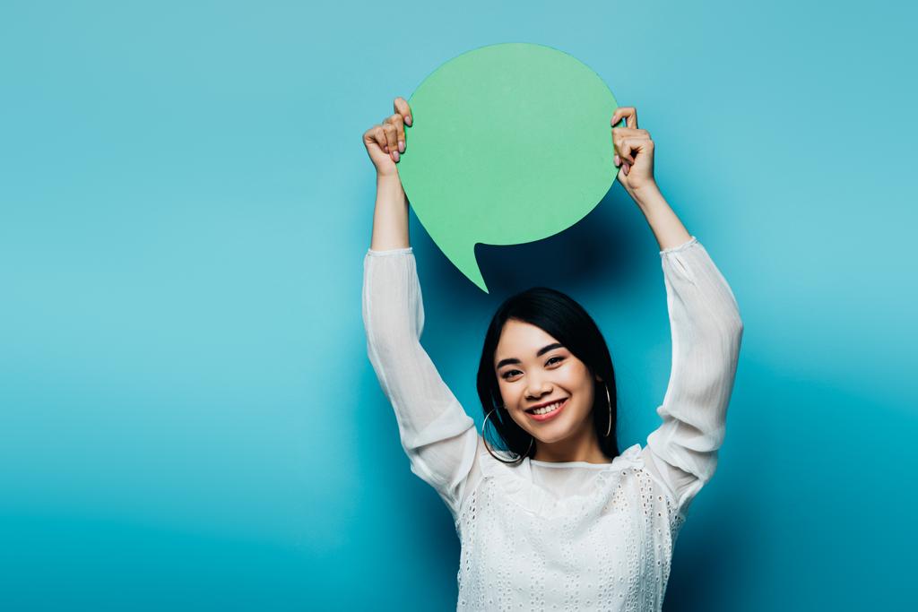 Gülümseyen esmer Asyalı kadın mavi arka planda yeşil konuşma balonu tutuyor. - Fotoğraf, Görsel