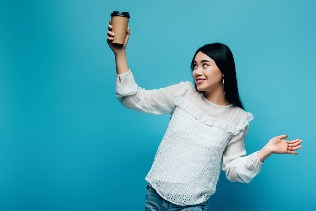 Gülümseyen esmer Asyalı kadın mavi arka planda kahve tutuyor. - Fotoğraf, Görsel