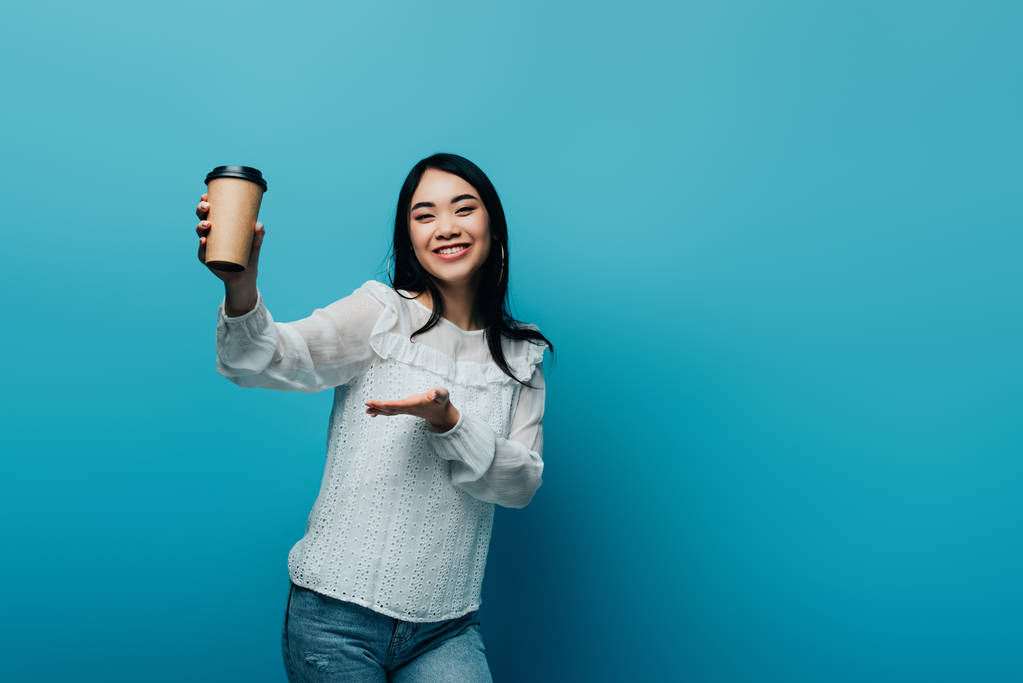 glimlachende brunette aziatische vrouw wijzend met de hand op koffie te gaan op blauwe achtergrond - Foto, afbeelding