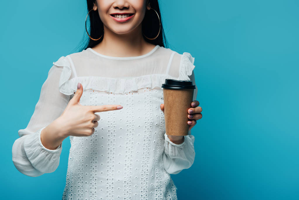 abgeschnittene Ansicht der lächelnden brünetten asiatischen Frau, die mit dem Finger auf Kaffee zeigt, um auf blauem Hintergrund zu gehen - Foto, Bild
