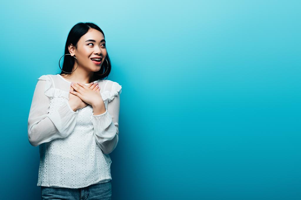 šťastný šokovaný bruneta asijské žena dívá pryč na modrém pozadí - Fotografie, Obrázek