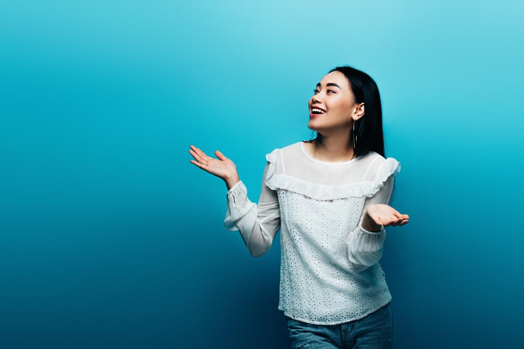 onnellinen brunette aasialainen nainen osoittaa kohauttaa ele sinisellä taustalla
 - Valokuva, kuva