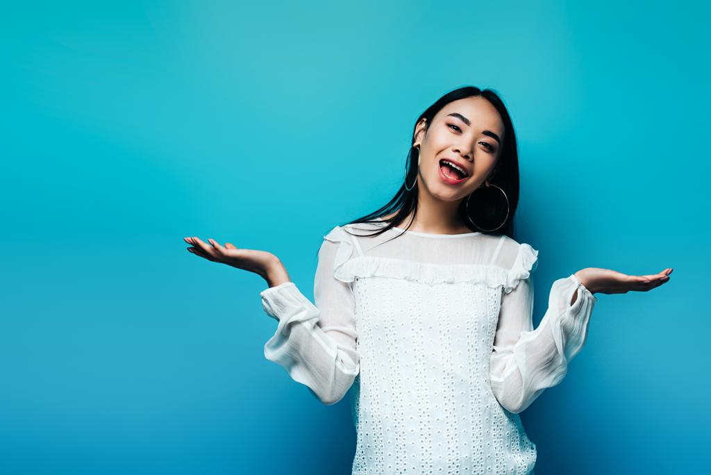 gelukkig brunette aziatische vrouw gebaren op blauw achtergrond - Foto, afbeelding