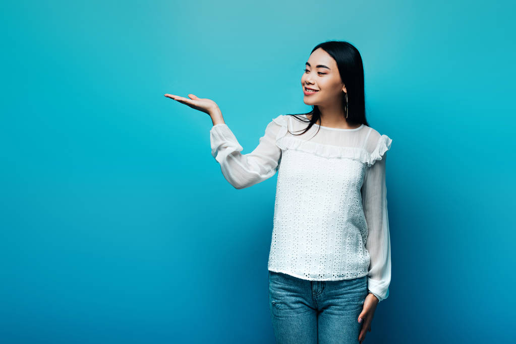 gelukkig brunette aziatische vrouw wijzend met de hand op blauwe achtergrond - Foto, afbeelding