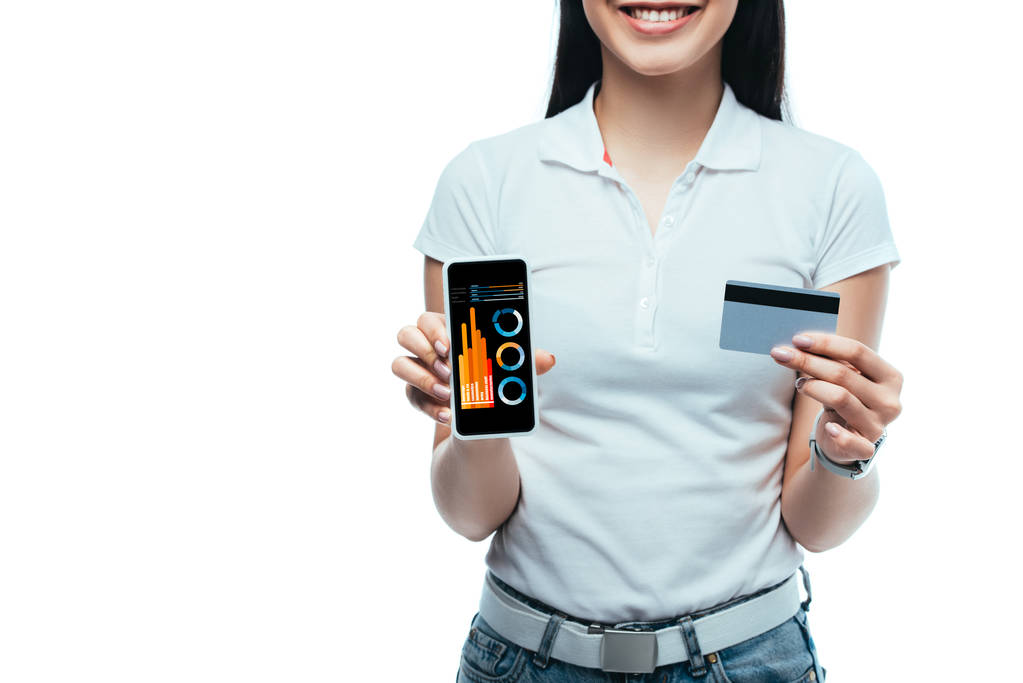 περικοπή άποψη του χαμογελαστού μελαχρινή ασιατική κορίτσι κρατώντας πιστωτική κάρτα και smartphone με επιχειρηματική εφαρμογή που απομονώνονται σε λευκό - Φωτογραφία, εικόνα