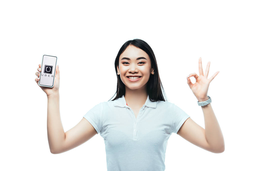 KYIV, UKRAINE - JULY 15, 2019: happy brunette asian woman holding smartphone with uber app and showing ok sign isolated on white - Photo, Image