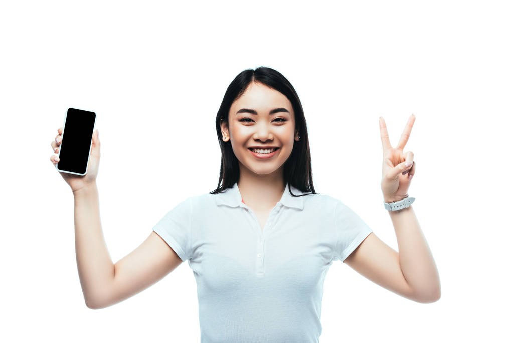 happy brunette asian woman holding smartphone with blank screen and showing peace sign isolated on white - Photo, Image