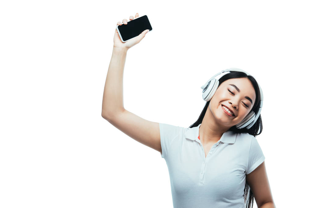 uśmiechnięta atrakcyjna azjatyckie dziewczyna w słuchawki gospodarstwa smartfon z pusty ekran odizolowany na biały - Zdjęcie, obraz