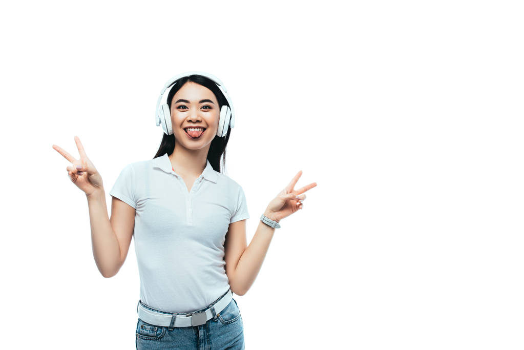 χαρούμενη ελκυστική Ασιάτισσα με ακουστικά που δείχνει σημάδια ειρήνης και γλώσσα απομονωμένα στα λευκά - Φωτογραφία, εικόνα