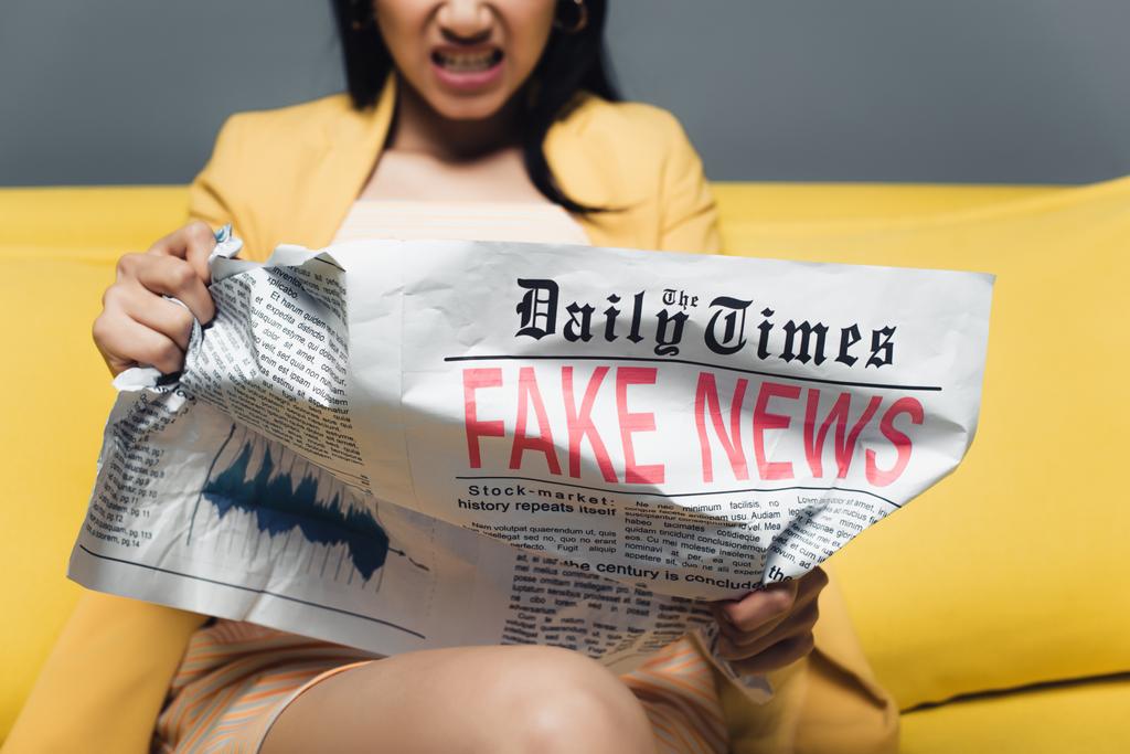 przycięty widok zły Azji businesswoman siedzi na żółtej kanapie i czytanie gazety z fałszywych wiadomości na szarym tle - Zdjęcie, obraz