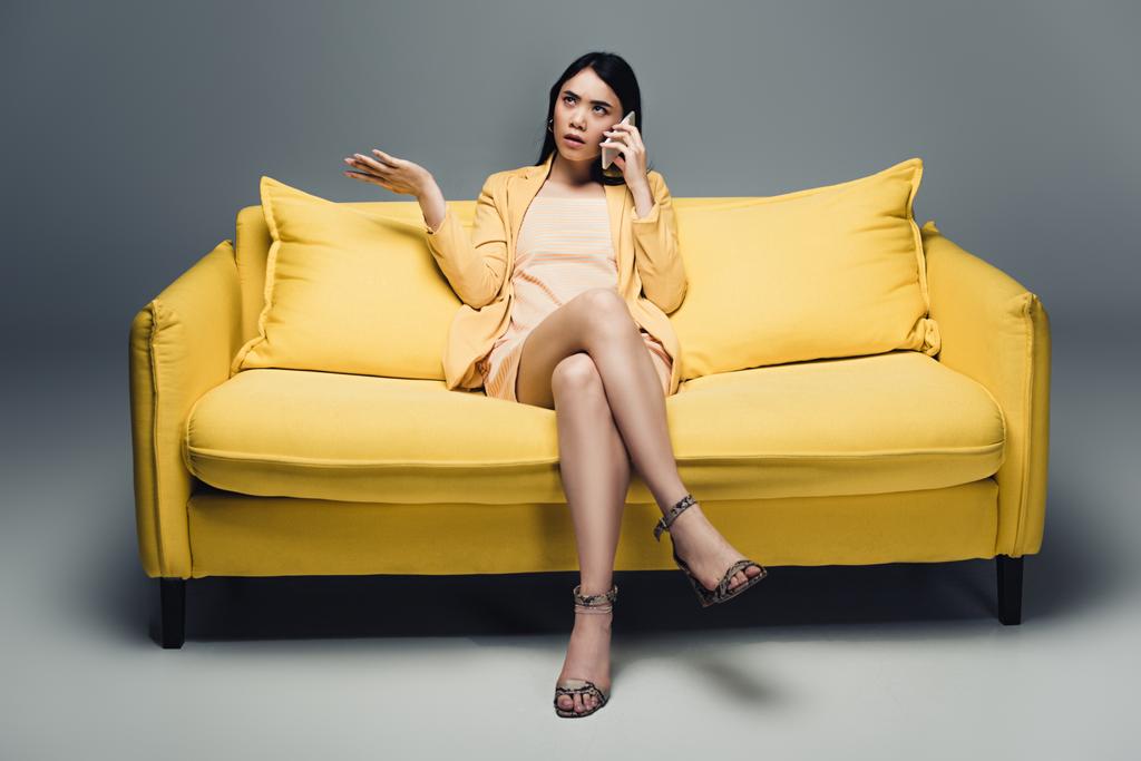 nespokojen asijské podnikatelka sedí na žluté pohovce se zkříženýma nohama a mluví na smartphone na šedém pozadí - Fotografie, Obrázek