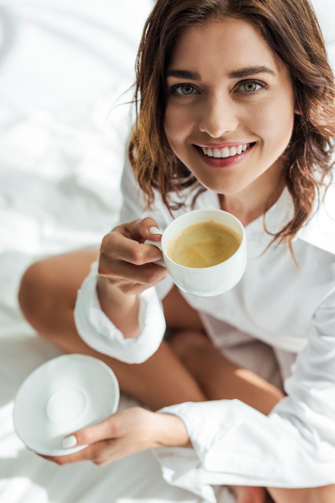 vista de ángulo alto de mujer atractiva en camisa blanca sonriendo y sosteniendo la taza por la mañana
  - Foto, imagen
