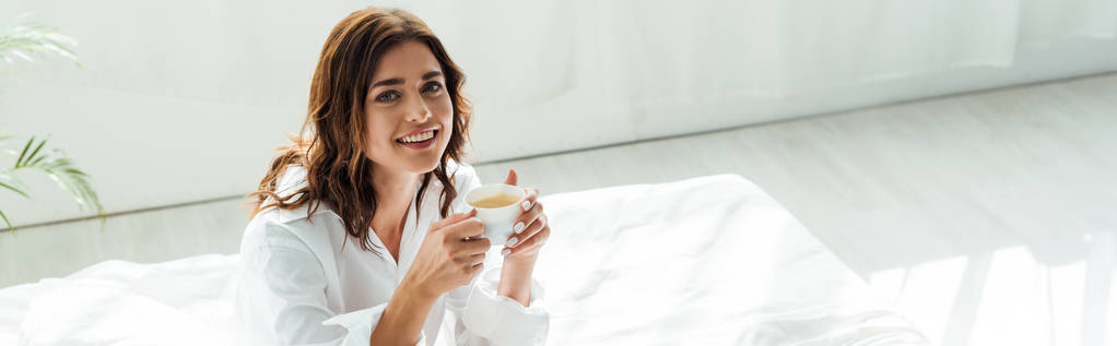 panoramiczne ujęcie atrakcyjnej kobiety w białej koszuli uśmiechającej się i trzymającej kubek rano  - Zdjęcie, obraz