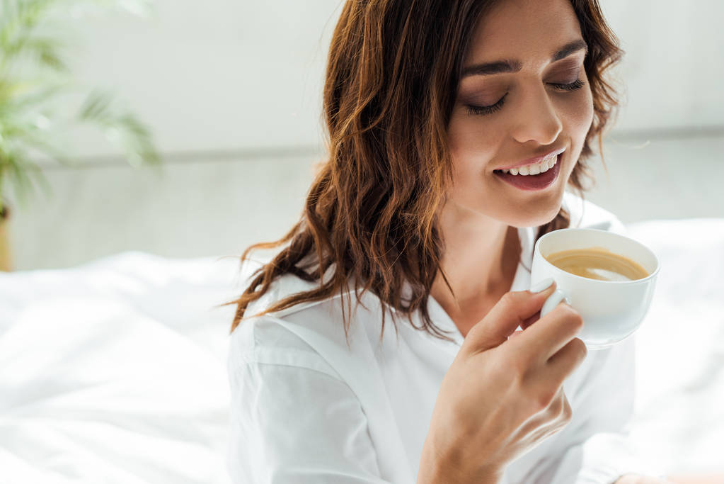 aantrekkelijke vrouw in wit shirt glimlachen en holding cup 's morgens  - Foto, afbeelding