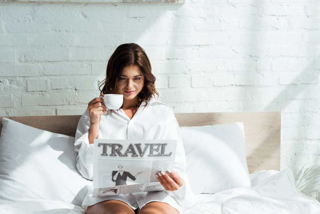 atractiva mujer en camisa blanca sosteniendo taza de café y leyendo viajes de periódico por la mañana
  - Foto, imagen